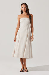 A woman is wearing a strapless, white, midi-length dress with a fitted bodice and a full, structured skirt. She has accessorized with gold earrings and a gold necklace, and is wearing black flip-flop style sandals.